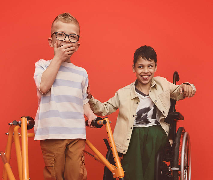 Two boys, one using a walking frame, and one in a wheelchair smile and laugh. The boy with the walking frame has his hand over his mouth.