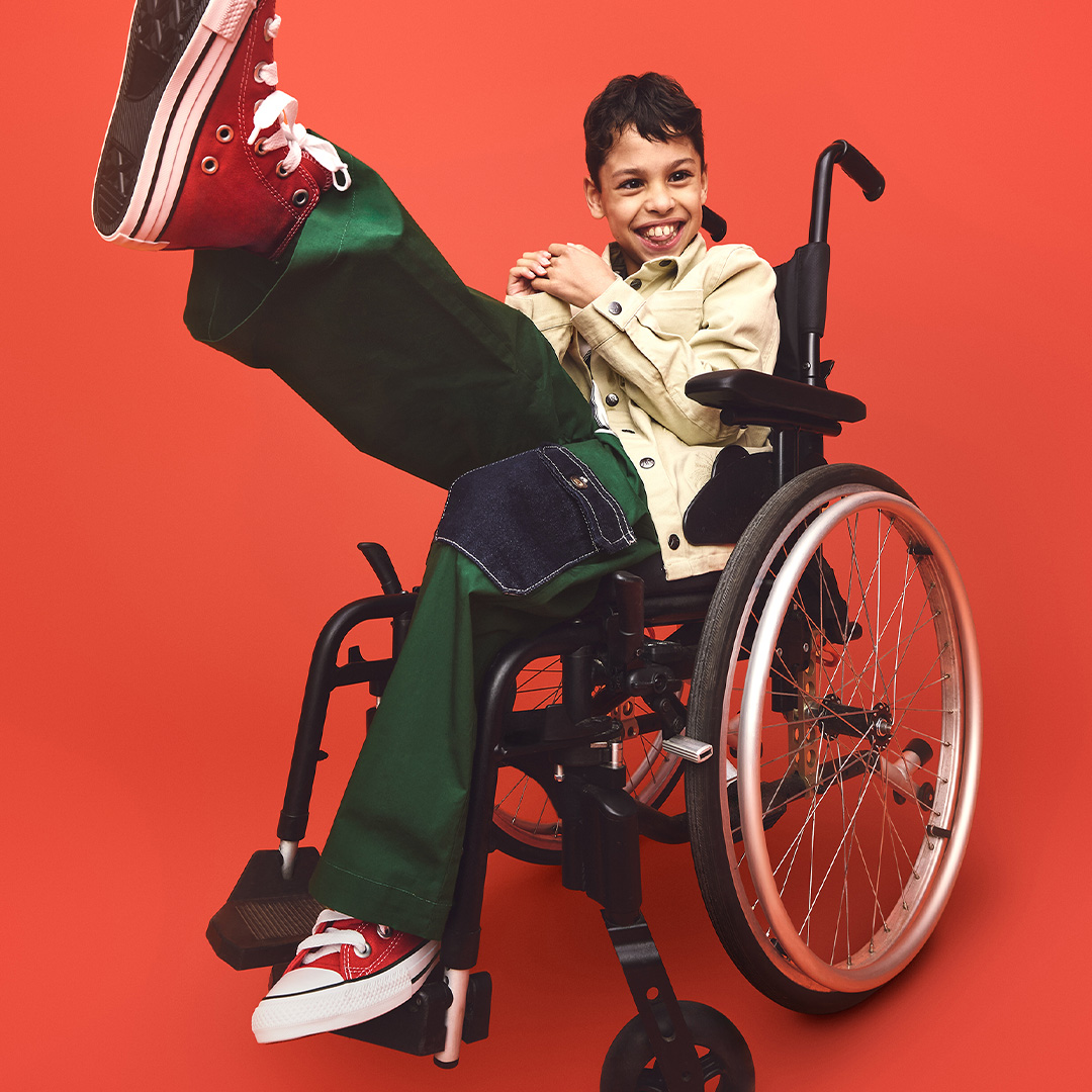 A boy in a wheelchair kicks up his left leg while wearing a happy expression.